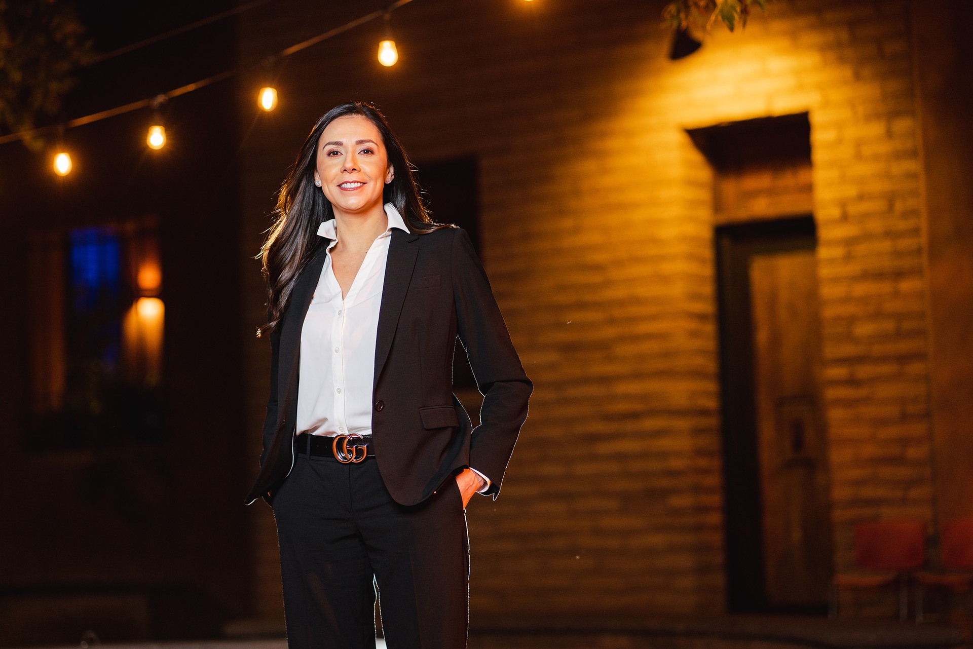 Woman in black blazer and white shirt standing in front of brick building with warm overhead lighting at night.
