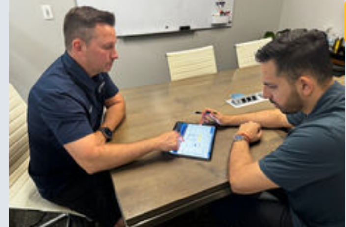 Two men sit at a table reviewing content on a tablet during a business discussion or training session.