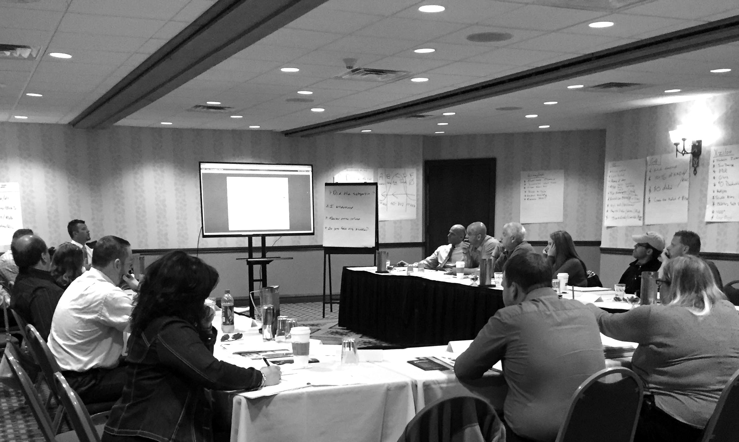 Professionals seated at tables during a conference room training session with a presenter at the front, display screen, and whiteboard notes visible.