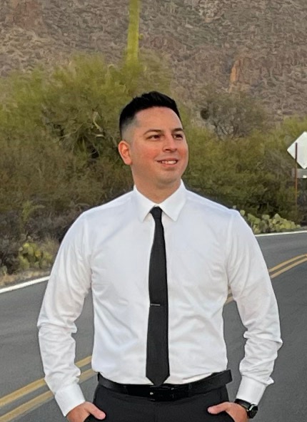 Man in white dress shirt and black tie standing on desert road with saguaro cactus in background.