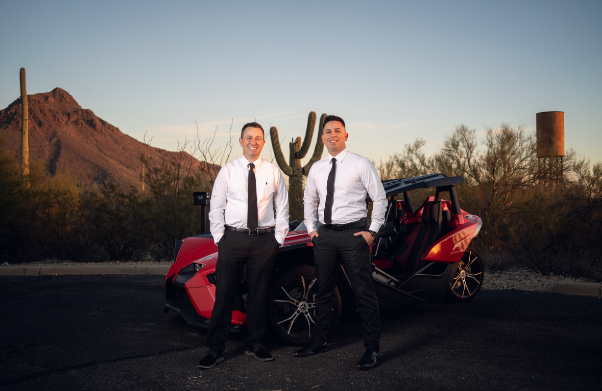 Two men in business attire standing between red recreational vehicles in a desert landscape with saguaro cacti.