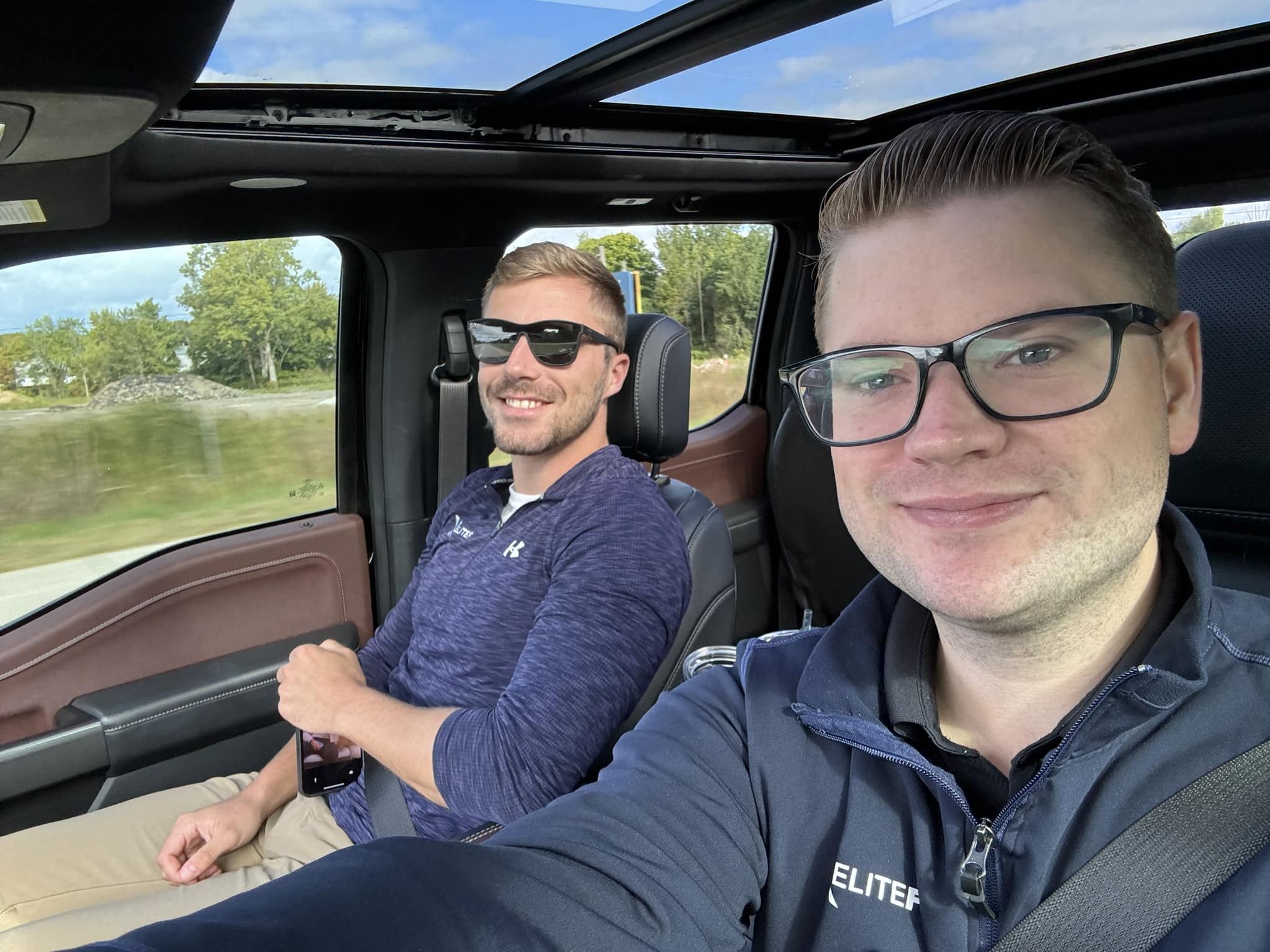 Two men sitting in a vehicle with open roof panels, smiling at camera with scenic lake and forest landscape visible through windows.
