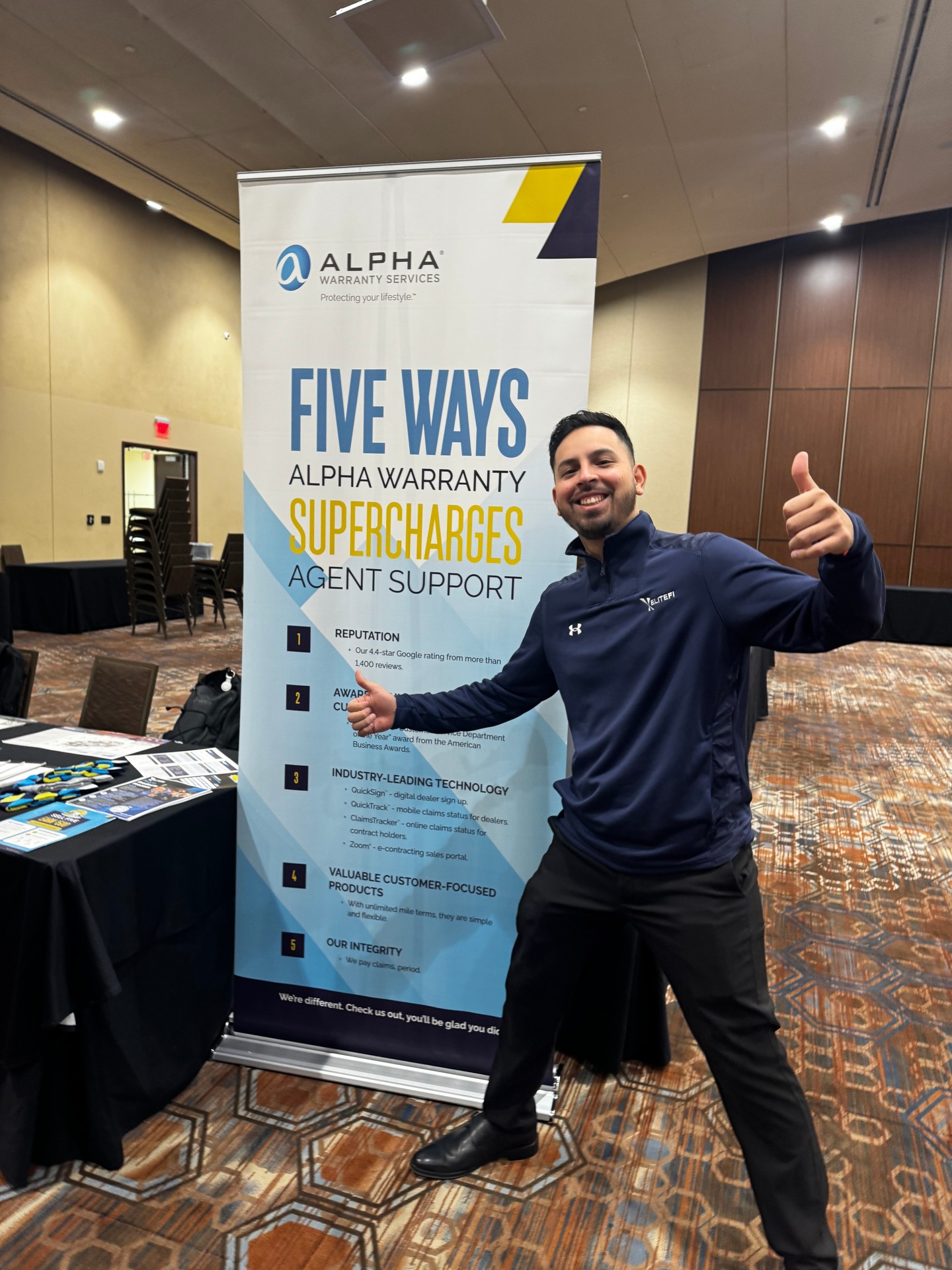 Man giving thumbs up at trade show booth with Alpha Warranty Services banner in convention center hallway.