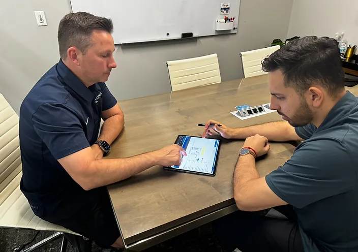 Two men sit at a wooden table reviewing content on a tablet during a one-on-one meeting in an office conference room.