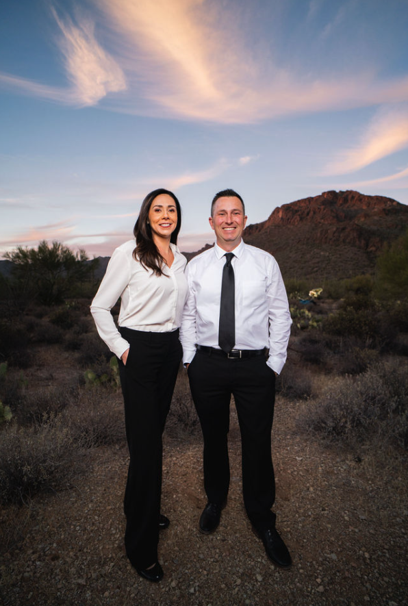 Two professionals in business attire standing in desert landscape with rocky hills and sunset sky behind them.