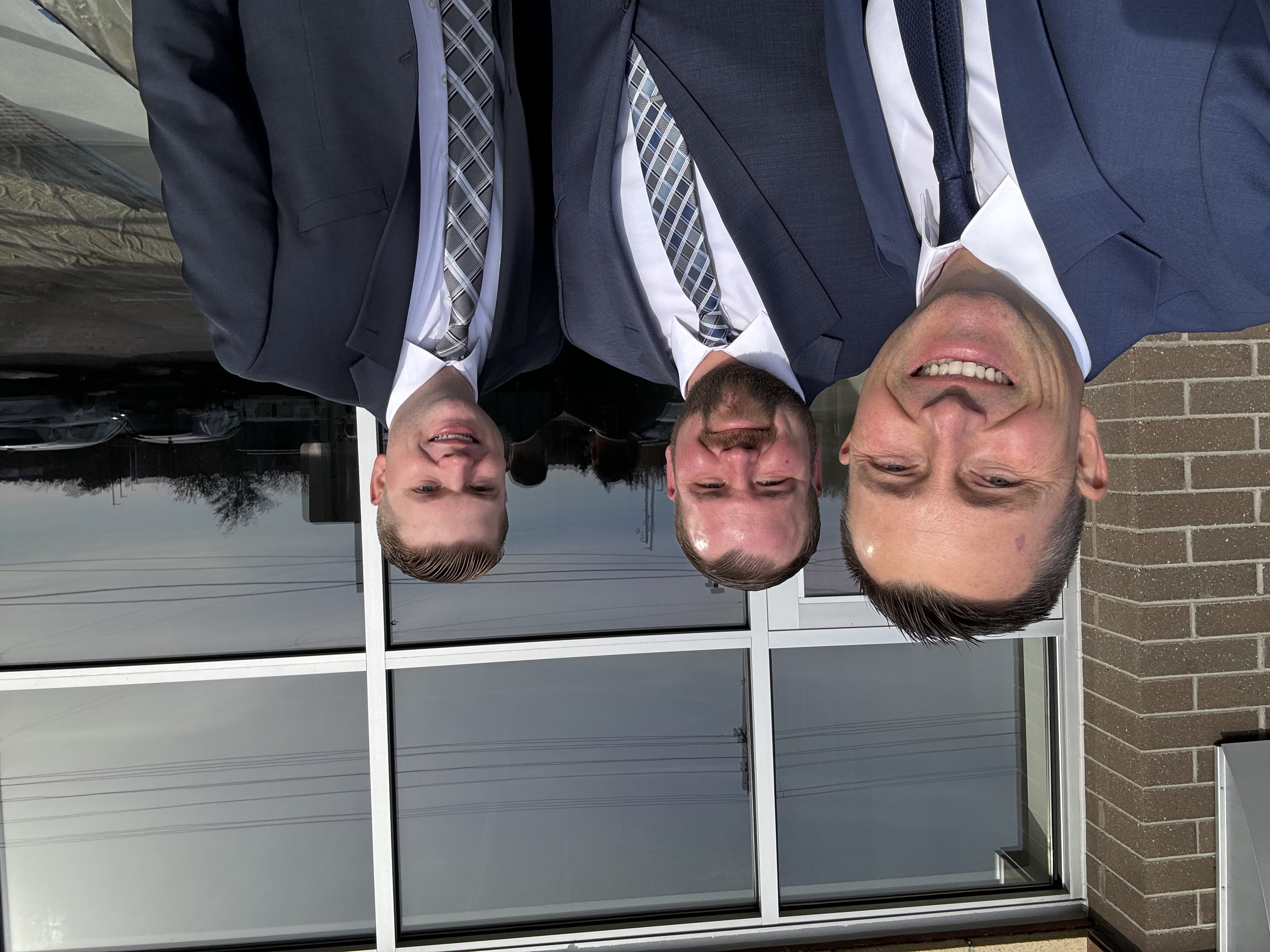 Three business professionals in dark suits and ties smiling at camera outside modern brick building with large windows.
