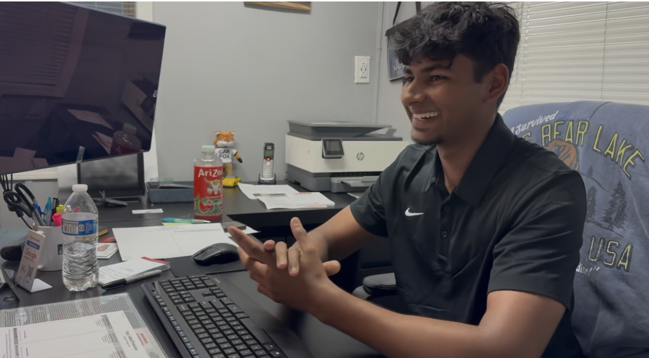 Young man in black Nike polo smiling while gesturing at desk with computer, printer, and office supplies in background.