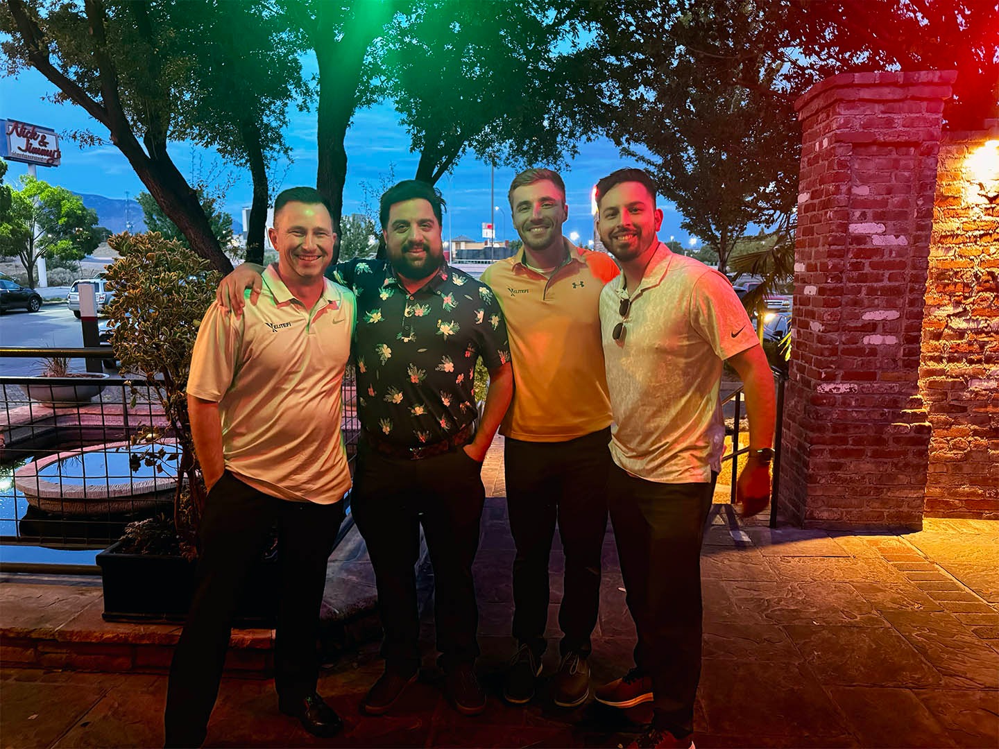 Four men standing together at dusk on a patio with brick building and large trees, smiling at camera in casual outdoor setting.