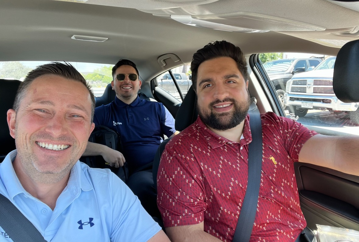 Three men smiling inside a car, parked lot visible through windows in background.