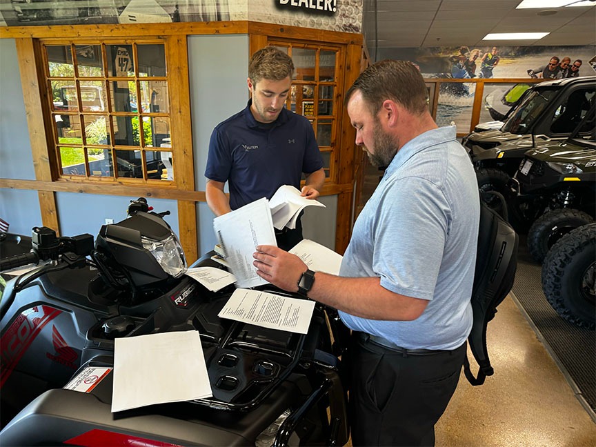 Two men reviewing paperwork at an ATV dealership showroom with vehicles and wooden display cabinets in the background.