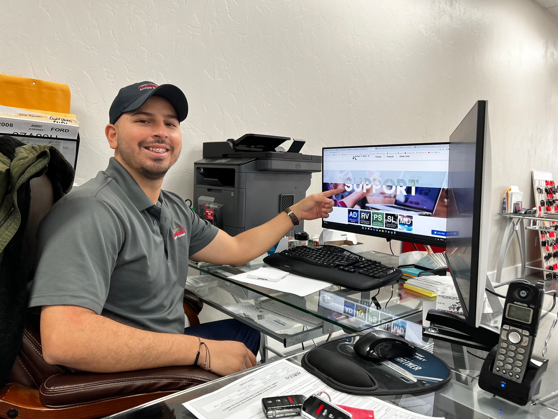 Man in gray polo and black cap smiling at desk while pointing at computer monitor displaying support website in automotive office.