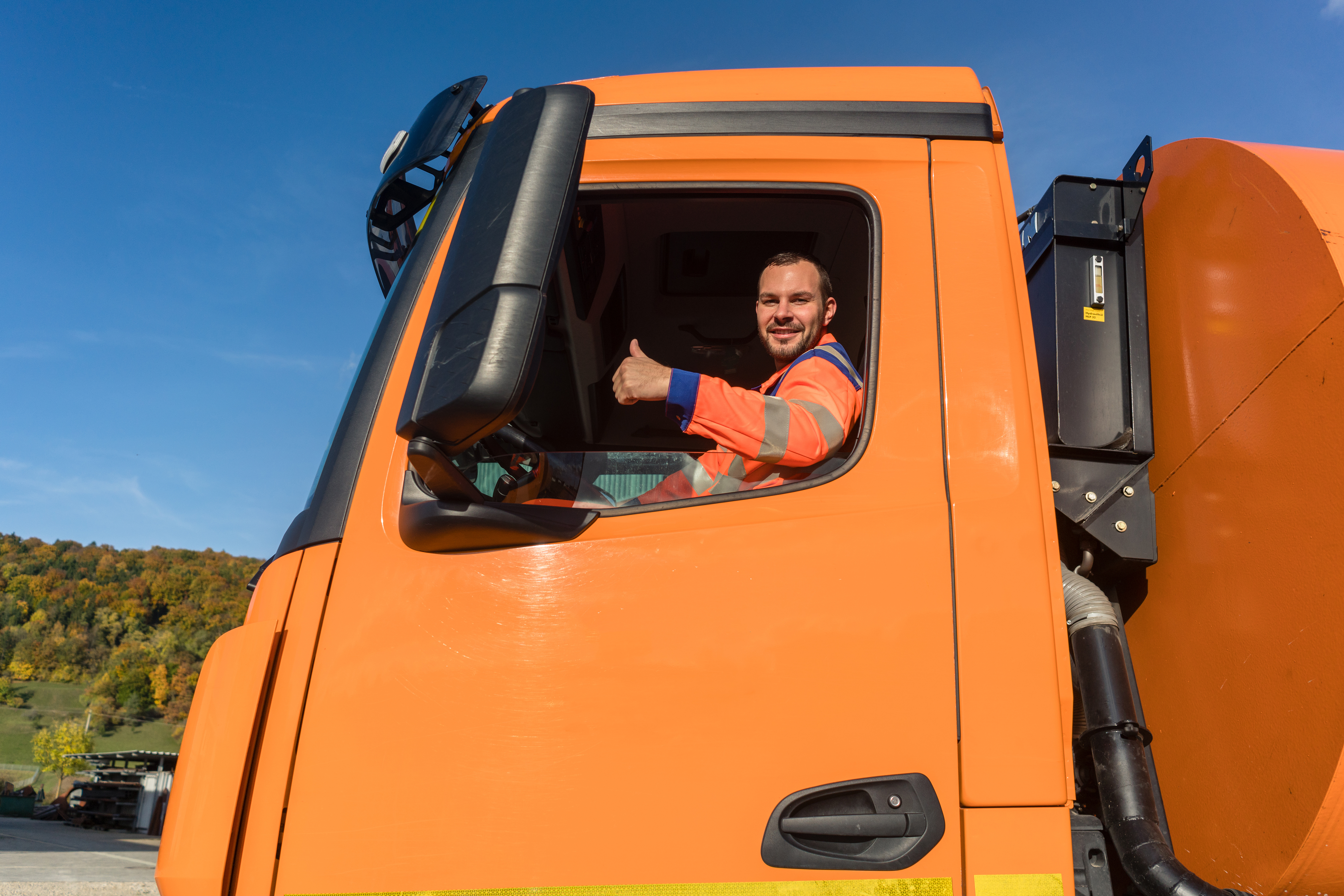 Truck driver in orange high-visibility vest giving thumbs up from bright orange tanker truck cab against clear blue sky.