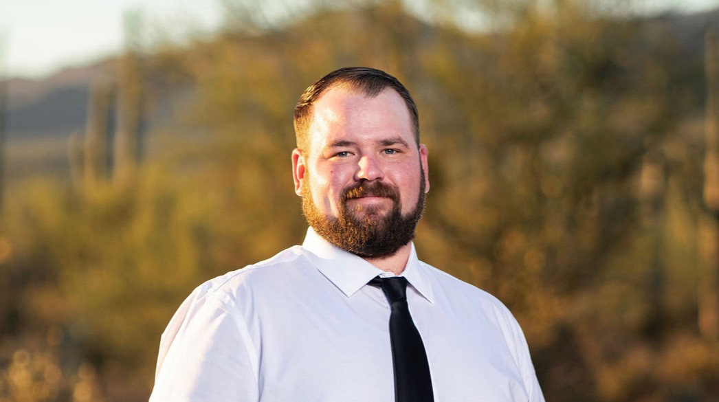 Man with beard wearing white shirt and black tie smiling at camera with autumn forest in background.
