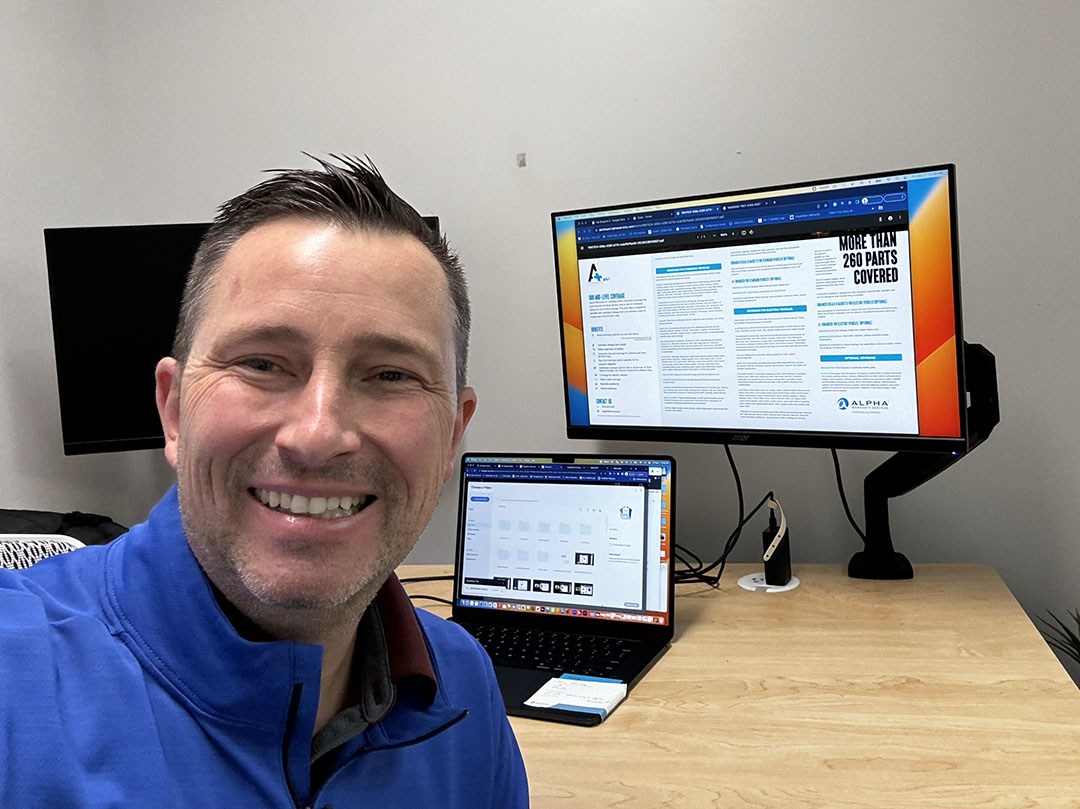 Man in blue shirt smiling at camera with multiple monitors displaying business presentations and data on wooden desk.