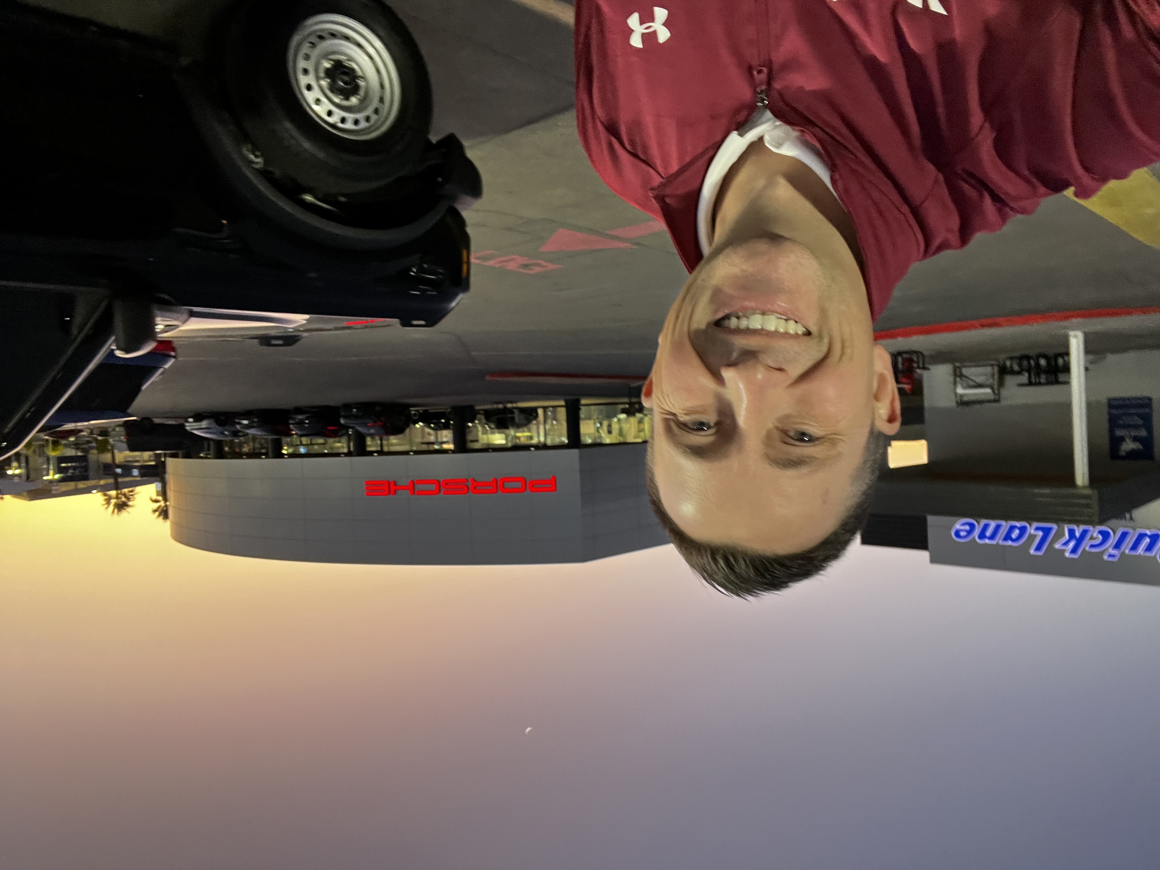 Man in maroon jacket smiling at Porsche dealership at sunset with crescent moon in sky