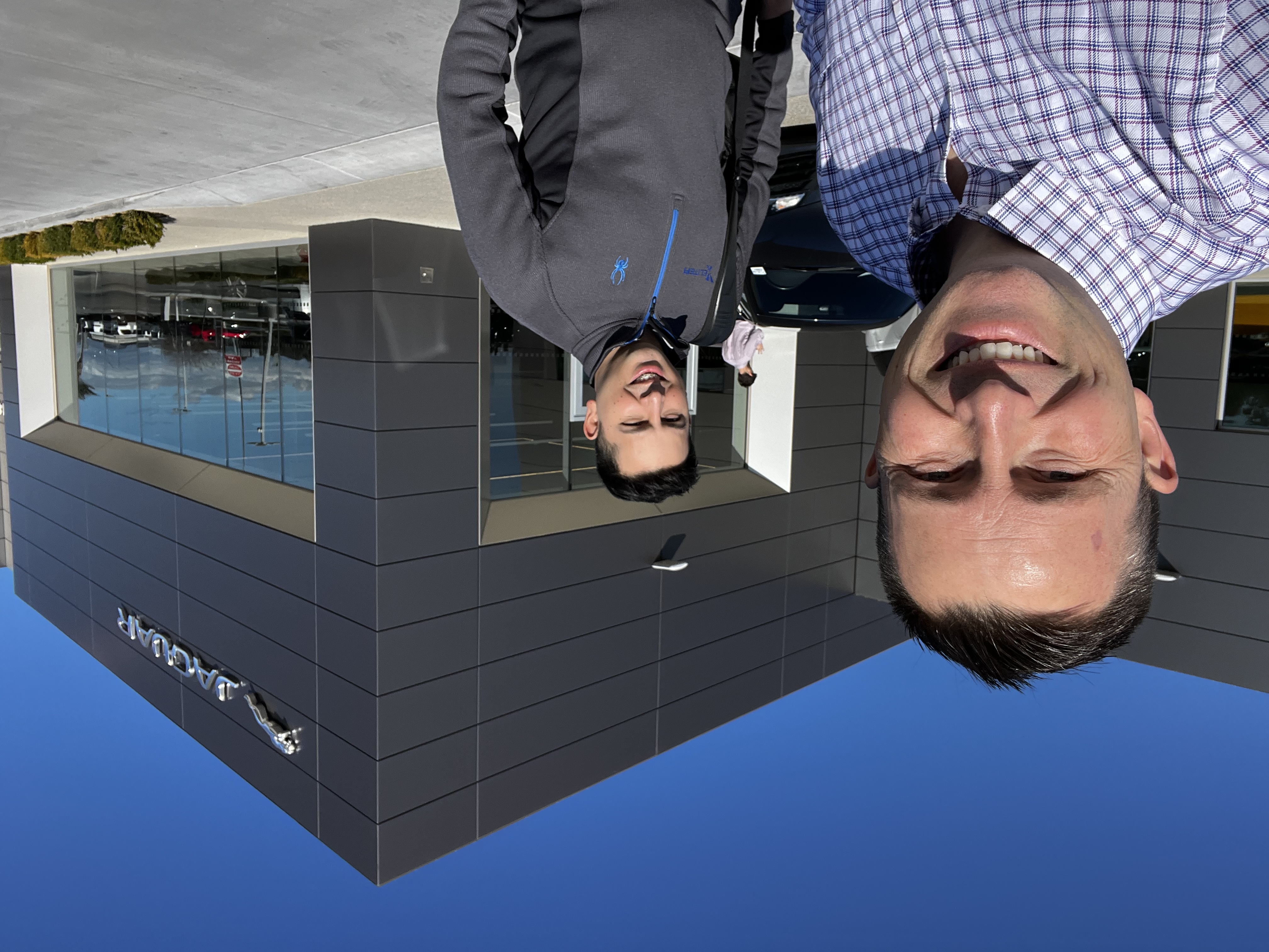 Two men smiling at a modern car dealership building with dark facade and large windows on a sunny day.