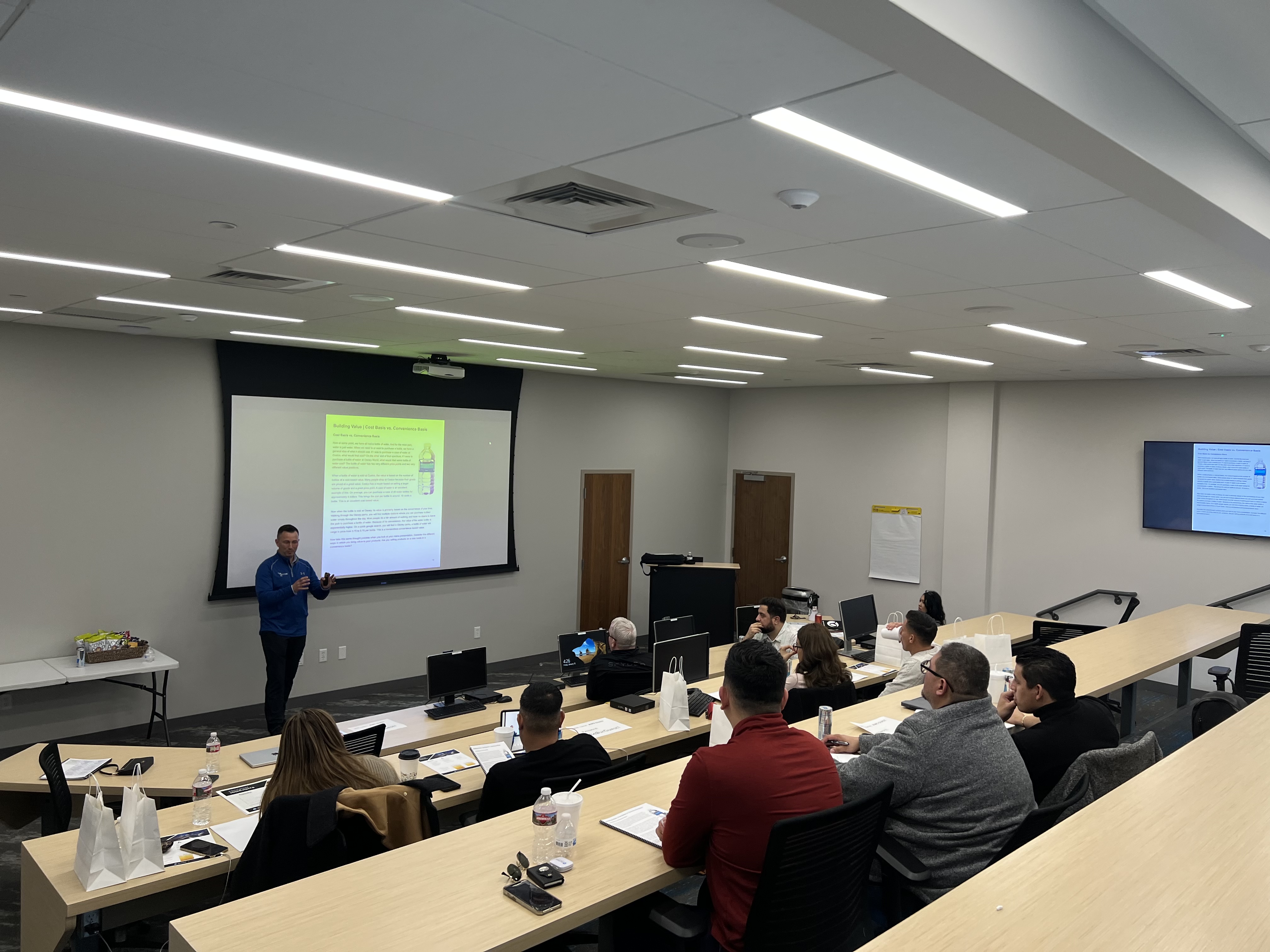 Presenter in blue jacket stands at front of lecture hall pointing to projected slide while students seated at tiered desks with laptops listen attentively.