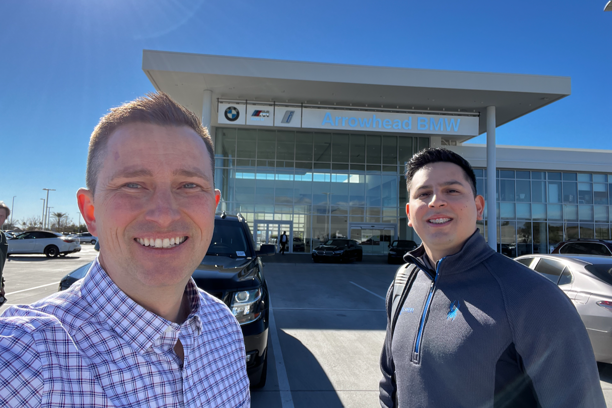 Two smiling men standing in front of Arrowhead BMW dealership on a sunny day with parked vehicles visible.