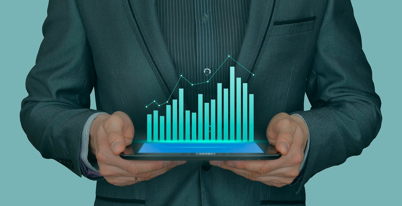Businessman holding tablet displaying holographic bar chart and upward trending line graph against teal background.