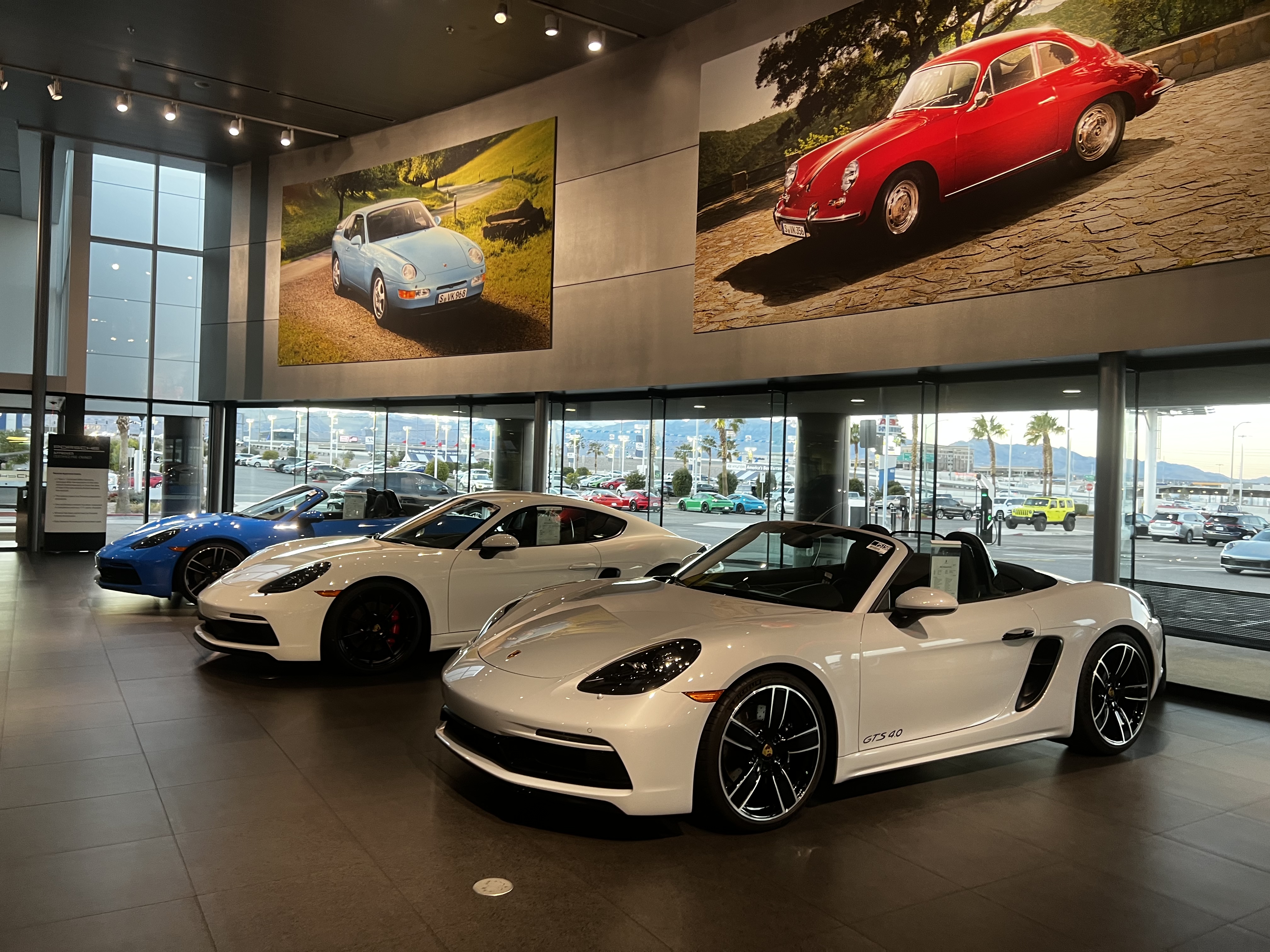 Modern sports cars displayed in a luxury dealership showroom with large windows overlooking a parking lot and mountains, vintage car photos on walls above.