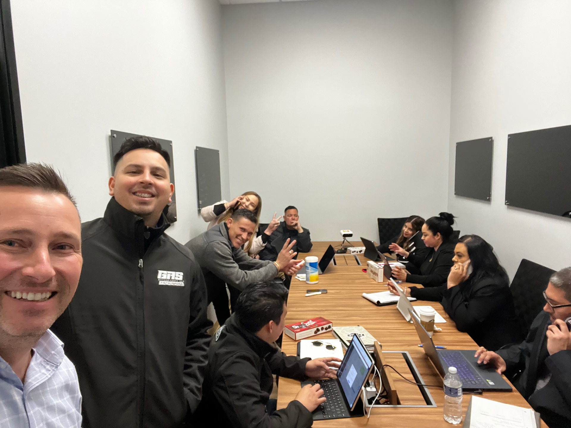 Group of professionals in business casual attire gathered around a wooden conference table with laptops, smiling and engaged in discussion in a modern office meeting room.