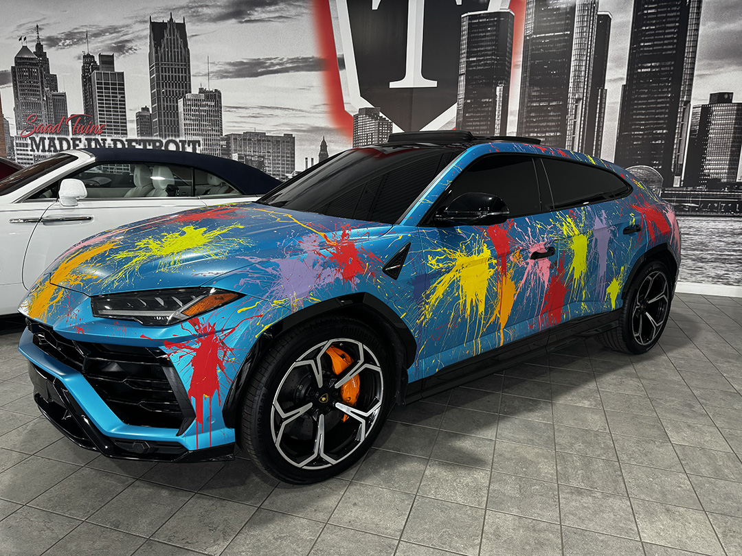 Blue sports car with vibrant red and yellow paint splatter art, parked in front of Detroit cityscape and "Made in Detroit" signage.