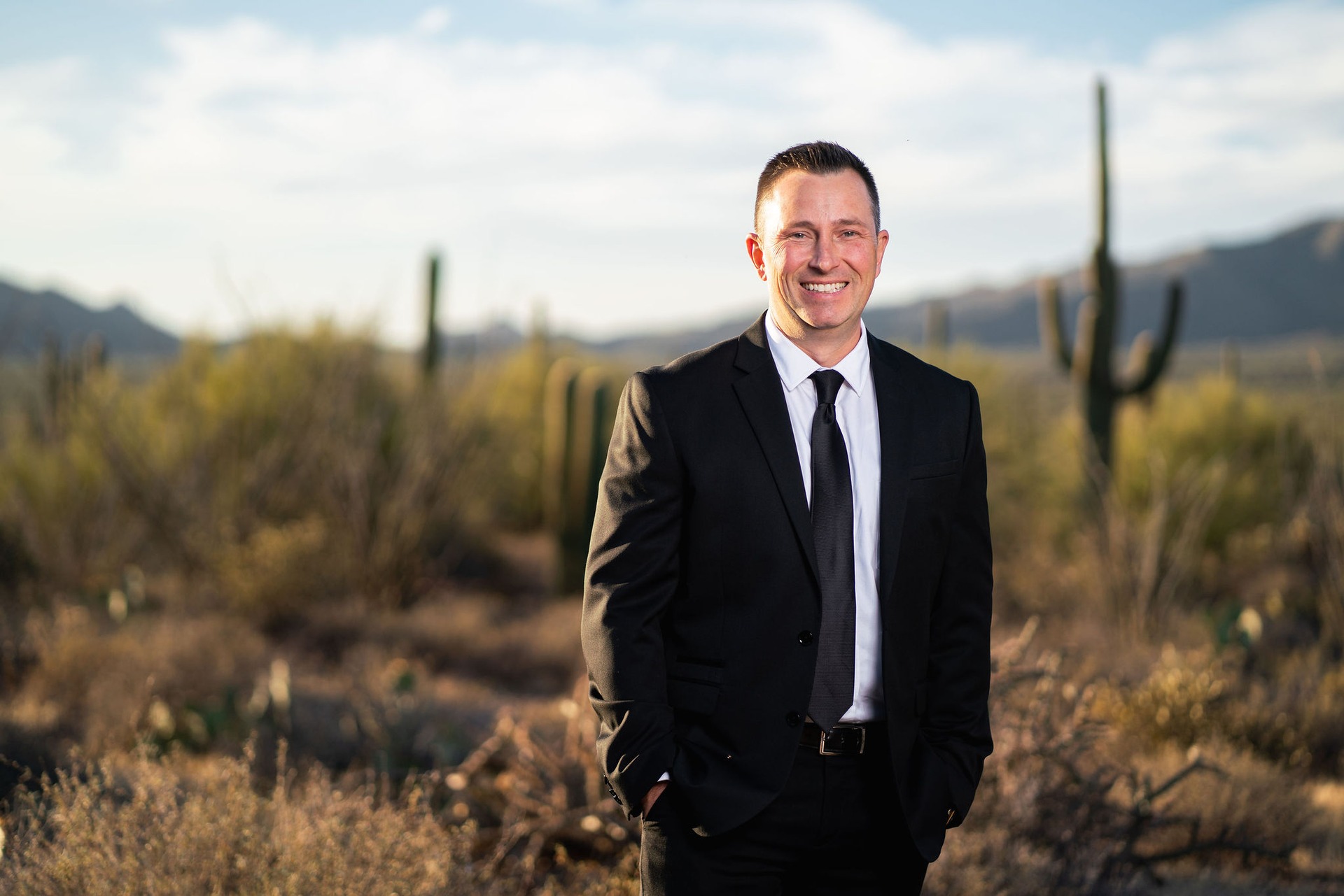 Smiling man in business suit and tie standing in desert landscape with saguaro cacti and mountains in background.