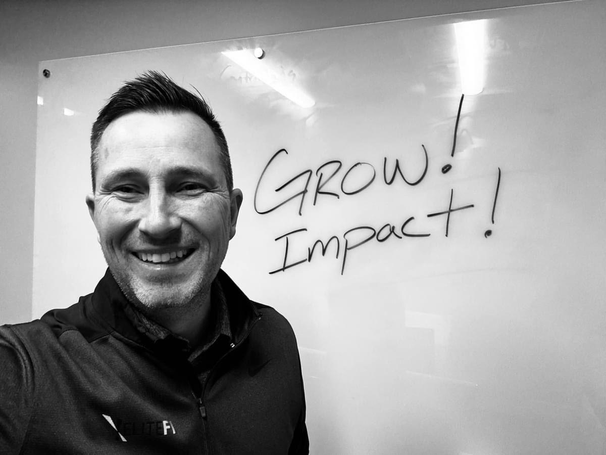 Smiling man in black jacket next to whiteboard with "GROW! IMPACT!" written on it.
