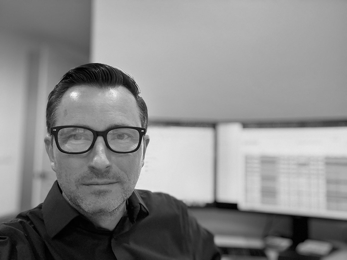 Man wearing glasses in modern office setting with computer monitors visible in background